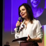 Foto de Daniela Grelin, mulher branca, com cabelos lisos na altura dos ombros. Ela segura um microfone em uma das mãos.