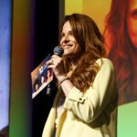 Foto de Michelle Lerni, mulher ruiva com cabelos longos e lisos. Ela fala ao microfone enquanto está em cima do palco do evento.