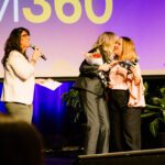 Foto de Karina Chaves falando ao microfone no palco, enquanto Lilian Rauld entrega um buquê de flores para a Margareth Goldenberg.