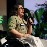 Foto de Carolina Ignarra, mulher cadeirante com cabelos lisos na altura dos ombros. Ela fala está no palco do evento e fala ao microfone.