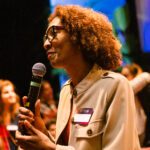 Foto de uma mulher negra de cabelos ruivos falando ao microfone.