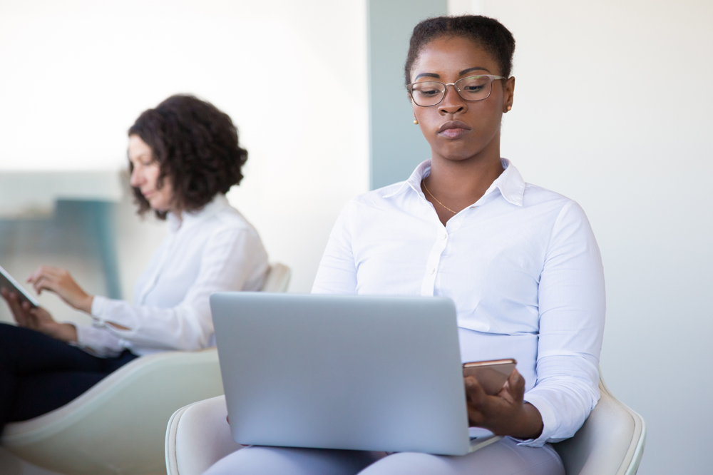 Foto de uma mulher negra sentada em uma poltrona digitando em um notebook que está em seu colo. No fundo, desfocado, há uma mulher branca de cabelos cacheados lendo.