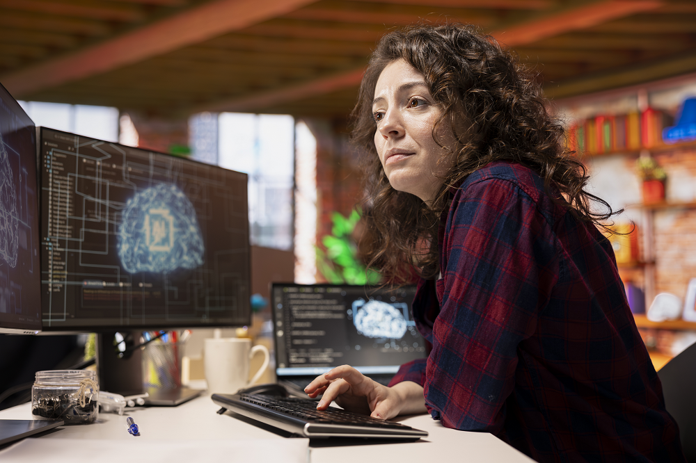 Foto de uma mulher sentada em um escritório. Sua mão está sobre o teclado do computador e na tela do monitor há a imagem de um cérebro.