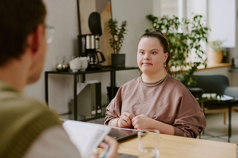 Foto de uma mulher com Síndrome de Down sentada de frente para outra pessoa em um escritório.