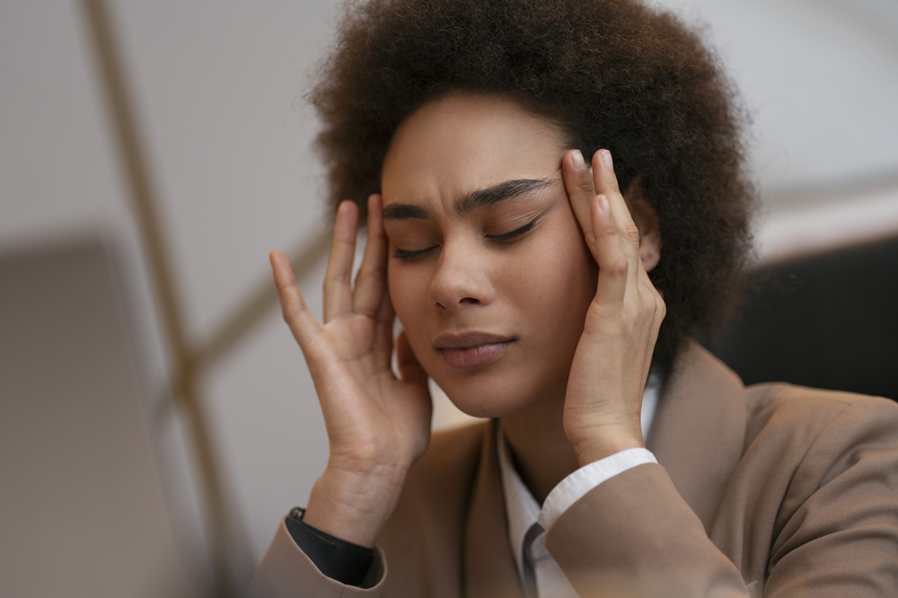Foto de uma mulher de cabelo curto e cacheado com os olhos fechados e as mãos nas têmporas.
