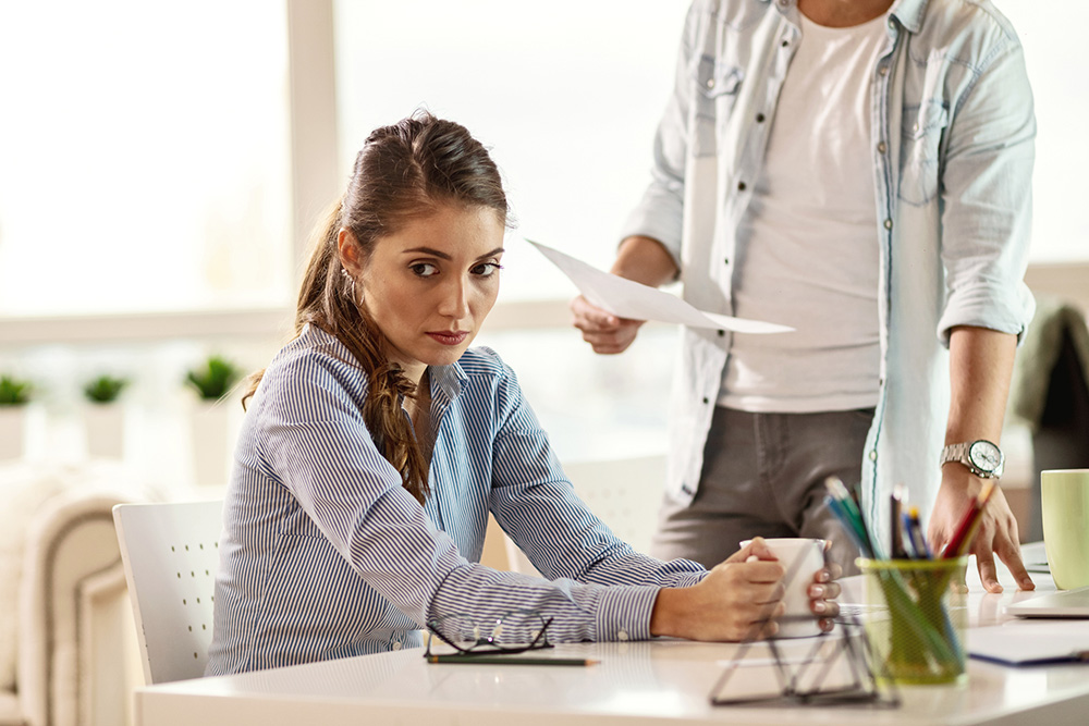 Foto de mulher de pele branca e cabelos castanhos claros usando roupa social sentada atrás de uma mesa de escritório, ela olha para o lado cabisbaixa. Ao lado dela, aparecendo apenas o dorso, há um homem em pé com um papel na mão.