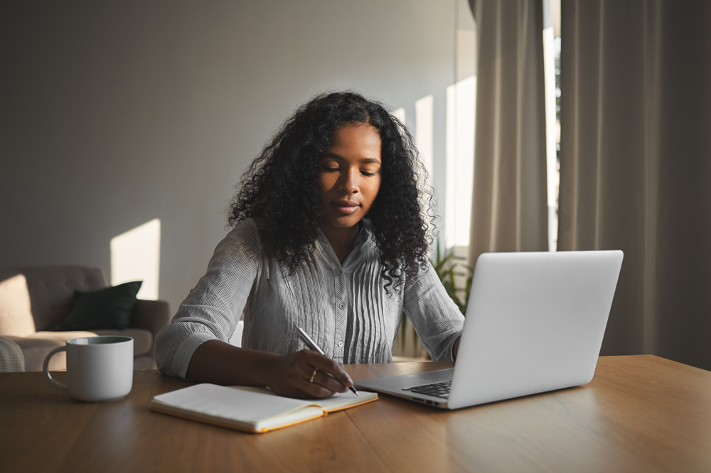 Foto de mulher negra jovem sentada à mesa com um laptop a sua frente.