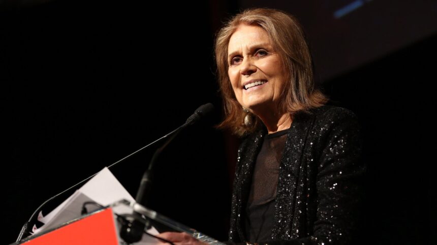 Foto de Gloria Steinem, mulher de pele branca, cabelos lisos e curtos na altura dos ombros. Ela fala ao microfone, veste uma roupa toda preta e sorri.